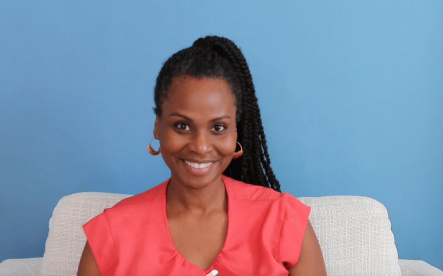 Sonia Thompson, a black woman with braids styled in a ponytail, sitting a white chair, with a blue background. She is wearing an orange short sleeved shirt and is smiling.