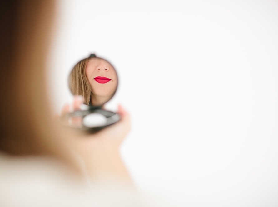 We can see a small round mirror in the style of a makeup holder, where the lower part of the face of a woman with red lips is reflected.