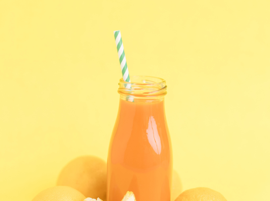 In the image we can see a container with orange juice and a straw. Around it segments of oranges and a background of the same color.