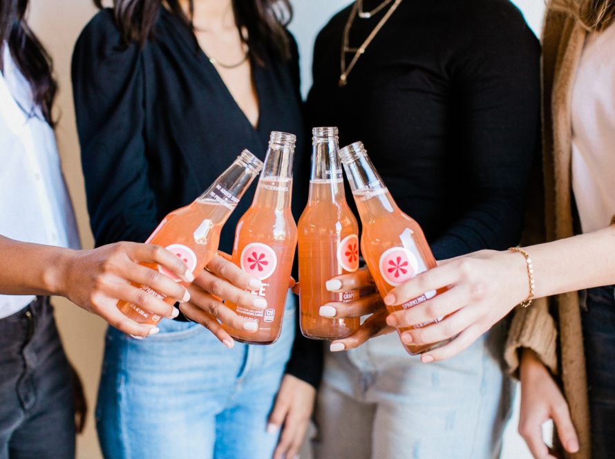 In the image, we can see a diverse group of women toasting their orange sodas.