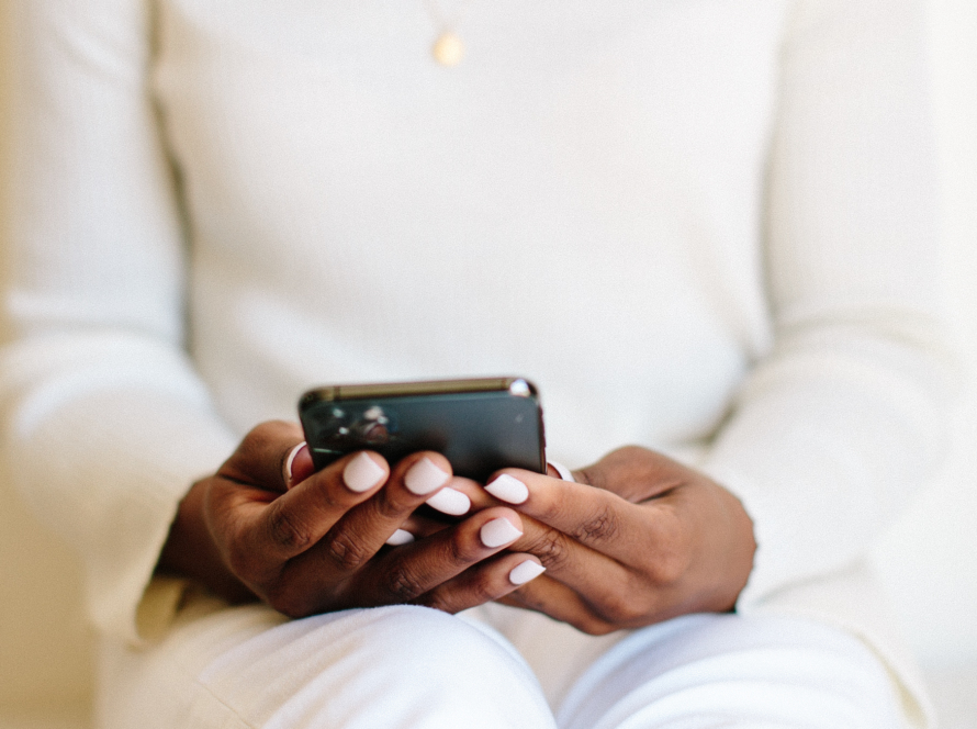 In the image, we can observe a black woman, dressed in white clothes, sitting down and holding a smartphone in her hands.