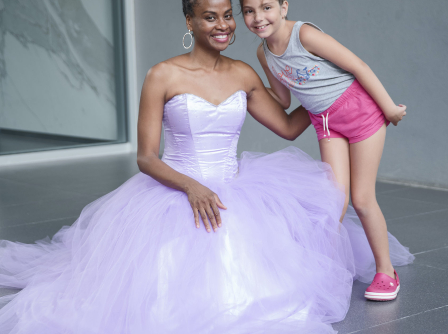Sonia Thompson, a Black-woman is wearing a purple princess dress and a tiara. Her hair is in a bun. She is kneeling down and smiling as she poses with a little girl. The white little girl about 7 years old is smiling, and wearing a grey tank top, pink shorts, and pink crocs. There is a grey background with grey tile floors.