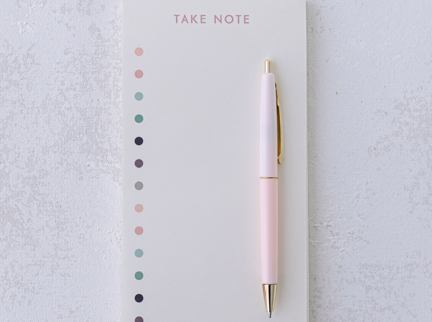 In the image we can see a notebook with white sheets, a pink pen and golden details.