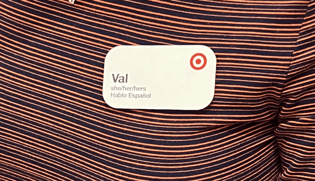 A black and orange horizontally striped shirt has a nametag with a Target red bullseye logo, the name Val with the pronouns she/her/hers underneath, and the phrase "Hablo Espanol" underneath the pronouns. This is an inclusive marketing example of a brand practicing "you've got options" category.