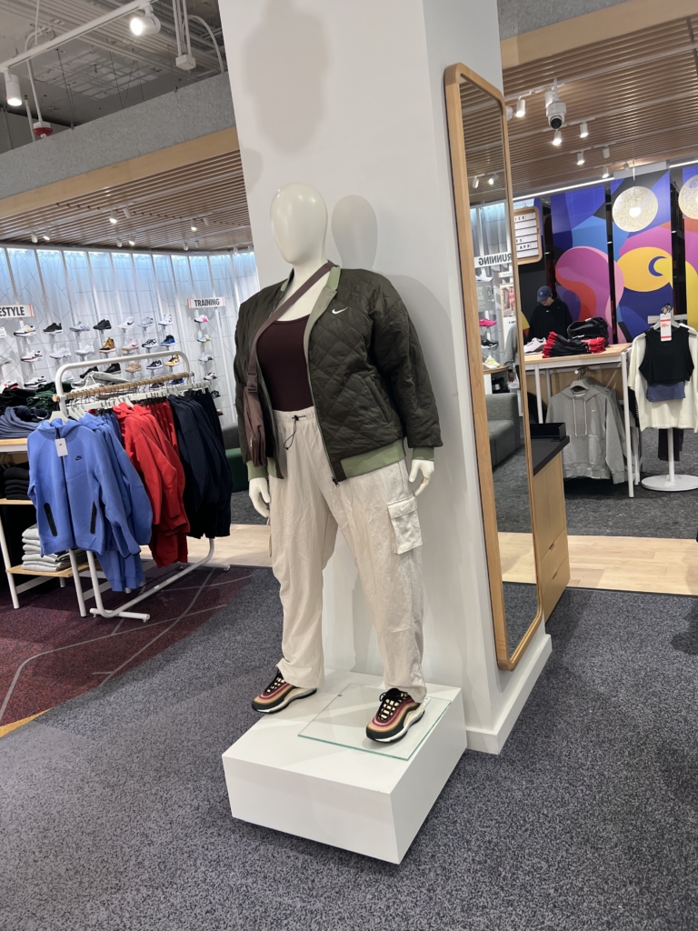 A pluz-sized female model is in the center of a sports retail store. It is an all white mannequin with a bald head. She is wearing cream color sweatpants, blue and orange tennis shoes, a brownish tanktop and a green jacket that is unzipped. She also has a brown purse hanging across her body. She is standing on a white pedastal.