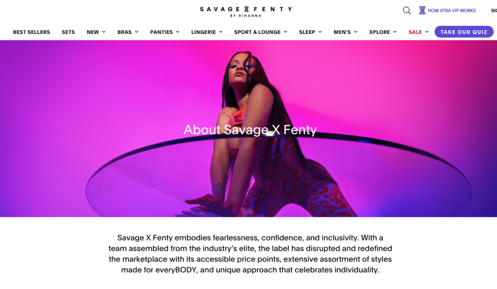 A screenshot of the Savage X Fenty by Rihanna website about page. The hero image has a hot pink and purple blended background with an image of Rihanna in the center. It appears as if she is kneeling on top of a round glass table and the camera is looking up at her from the bottom. Her hair is in long waist length braids, and she is wearing a two piece red and purple paisley jumper set. The top is a halter, and the bottom look like leggings. The copy reads, "Savage X Fenty embodies fearlessness, confidence, and inclusivity. With a team assembled from the industry's elite, the label has disrupted and redefined the marketplace with its accessible price points, extensive assortment of styles made for everyBODY, and unique approach that celebrates individuality."