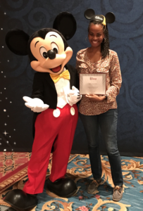 Sonia Thompson, a Black woman wearing blue jeans, and a flowered orange, red, and white shirt with long sleeves - with blue loafers. Her hair is in twists and are in a ponytail to the side. She's wearing a Mickey Mouse ears hat with a graduation tassle on it, and is holding a certificate. Next to her on the left is Mickey Mouse -- a large happy mouse, wearing a black jacket, red pants, and large black shoes. He's got white coves on his hand. There's a blue background in the back, and ugly floral carpet they are standing on.