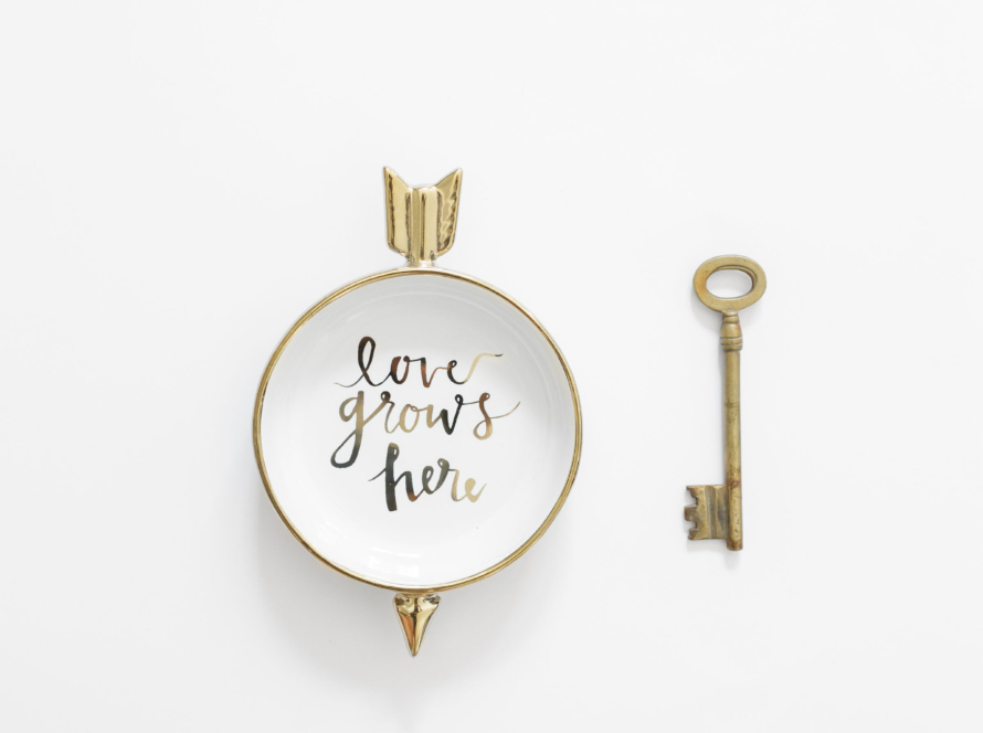 In the image we can see an old-style key and a composition ornament in which a circle with a white background and golden edges contains the phrase "Love Grows Here" and is crossed by an arrow from behind.