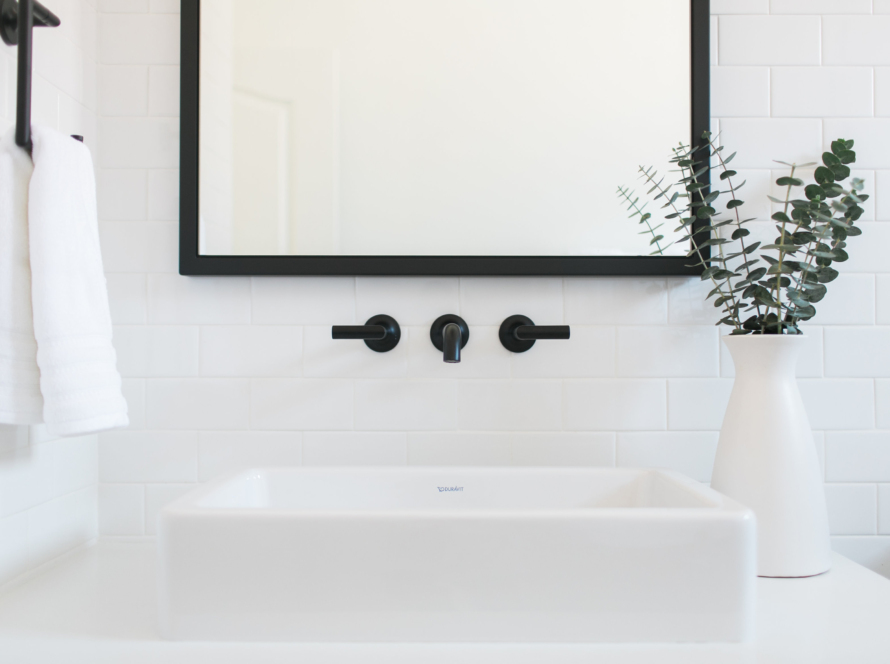 In the image, we can see a sector of an immaculate, clean white bathroom, with a vanity, white walls, and towels, taps, and a mirror frame in black.