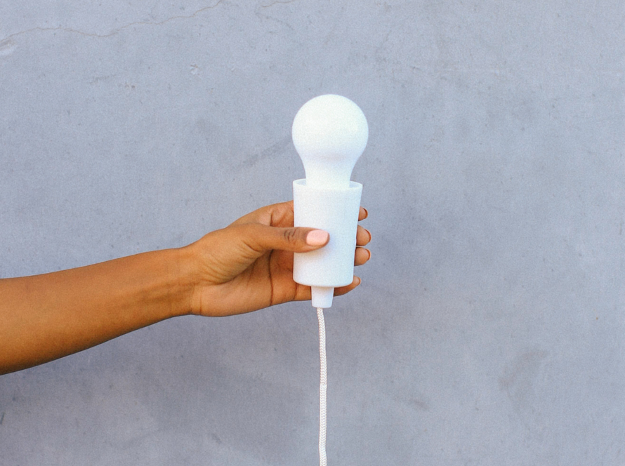 In the image we can see the arm of a black woman holding a lamp holder with its white lamp on a pastel blue background