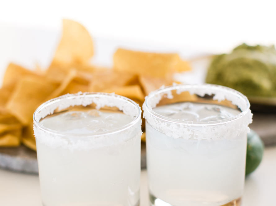 In the image we can see two glasses of tequila, with snacks and two limes in front of them.