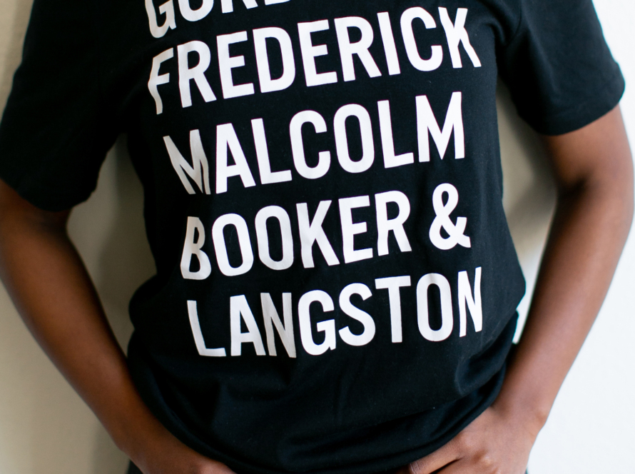 An image of a Black woman wearing a black t-shirt with these names written in white and in all caps: Gordon, Frederick, Malcolm, Booker, & Langston